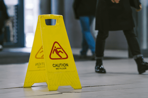 A caution sign warns of potential tripping and slipping hazards as people walk buy. You can only see the legs of the pedestrians.