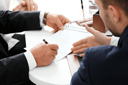 A business lawyer instructs his client on different laws regarding commercial litigation. The business owner is signing paperwork as the lawyer signals with his hand.