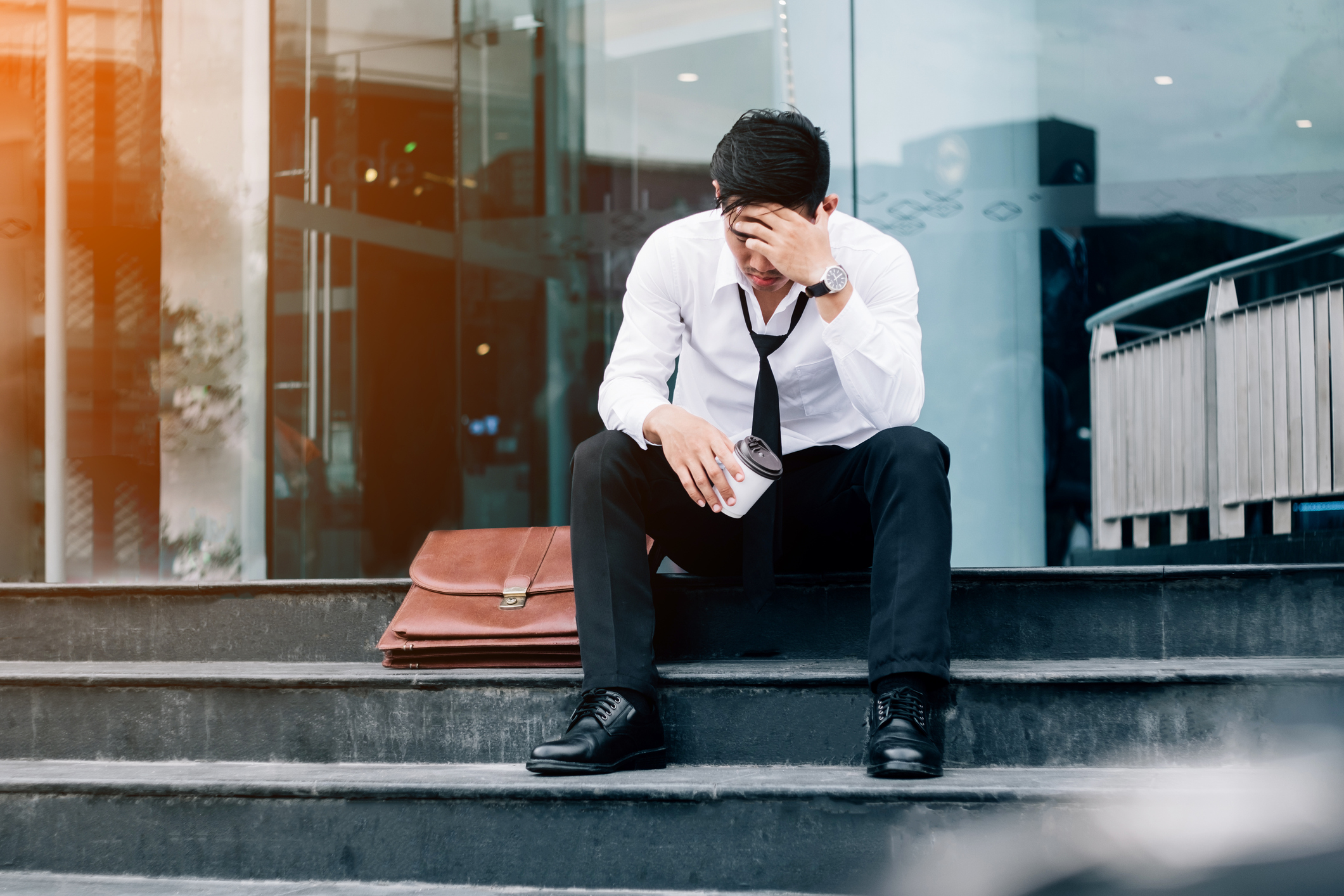 A stressed man in a shirt and tie sits on steps with his head in his hands.