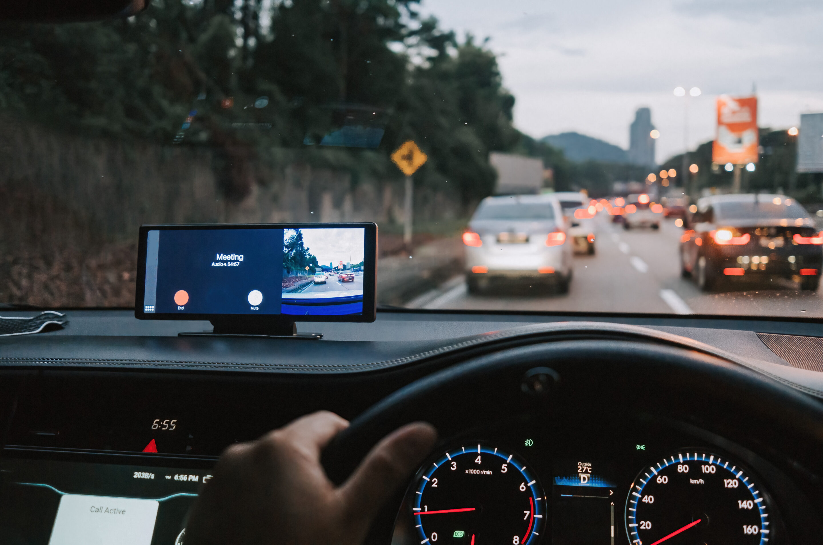 A phone on a car dashboard shows a video meeting while the car is in traffic.