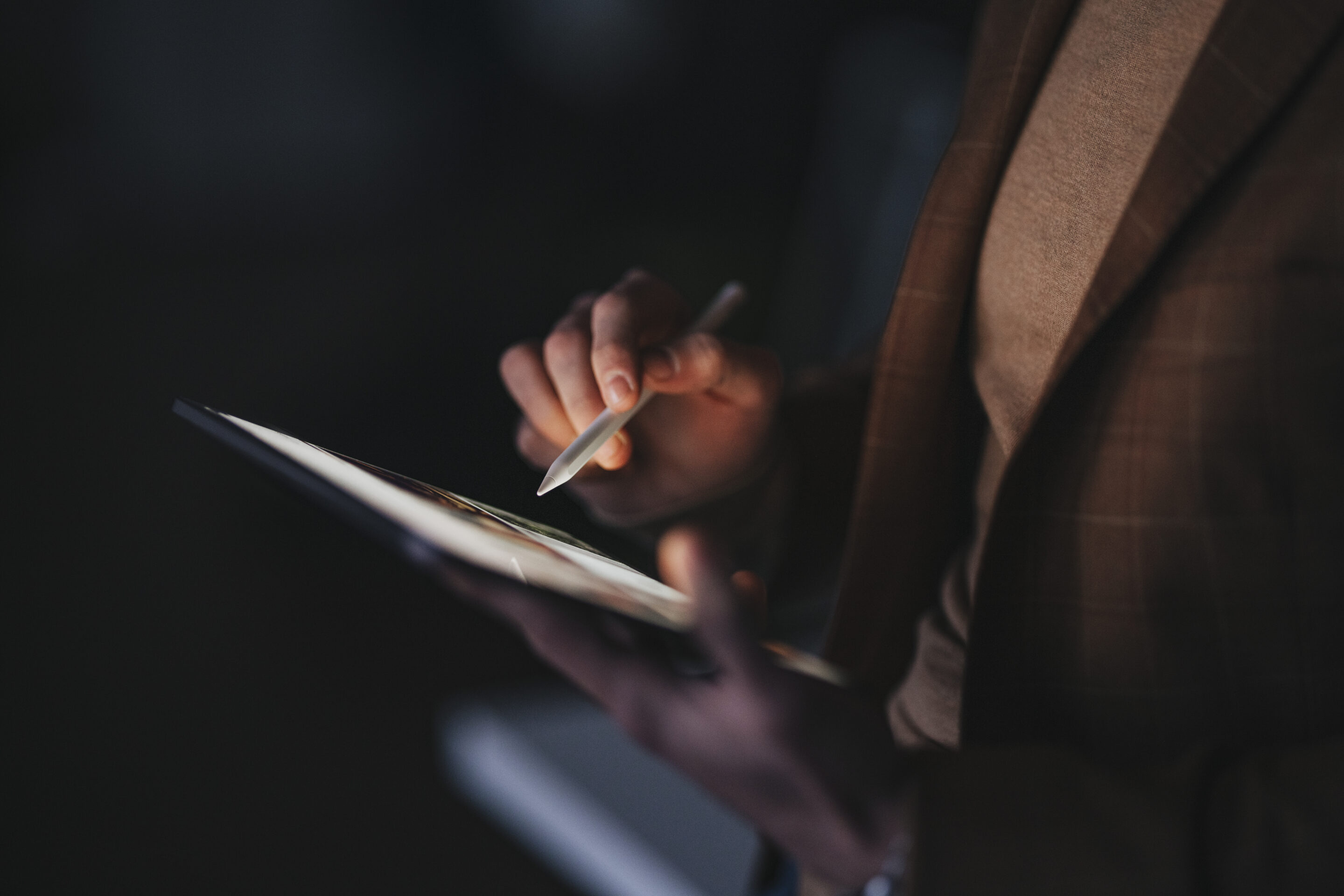 A person's hands use a stylus on a tablet screen in a dimly lit room.