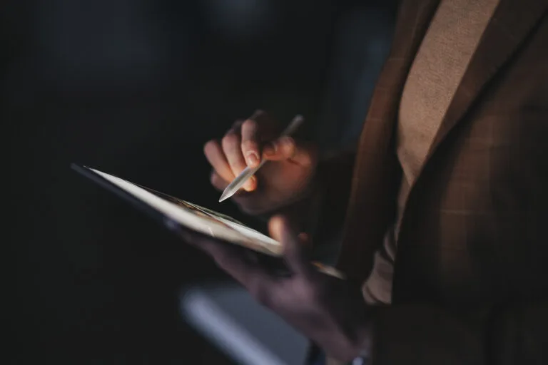 A person's hands use a stylus on a tablet screen in a dimly lit room.